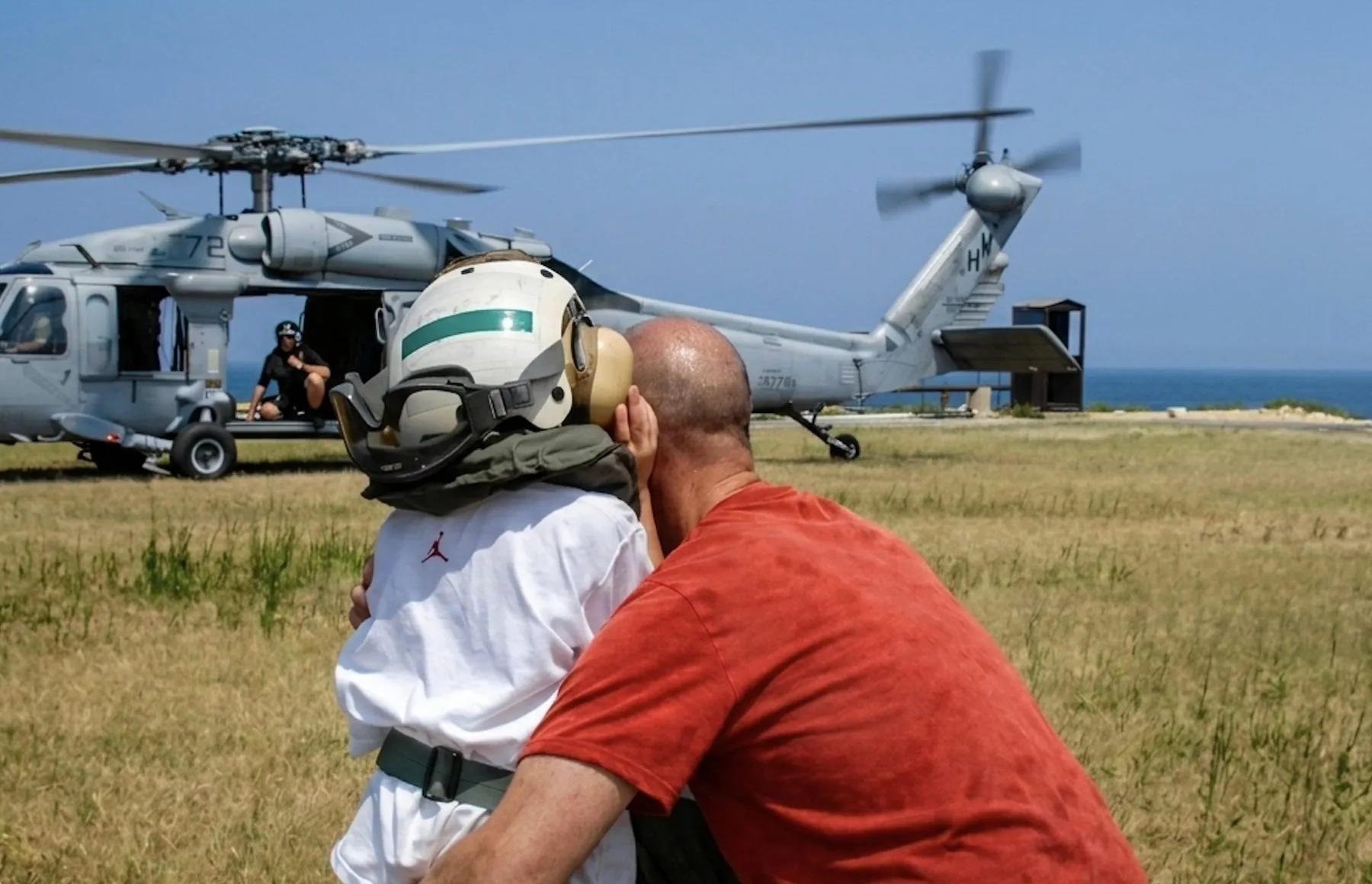 An evacuee hugs a child wearing a flight helmet beside a U.S. Navy helicopter, the sea behind them