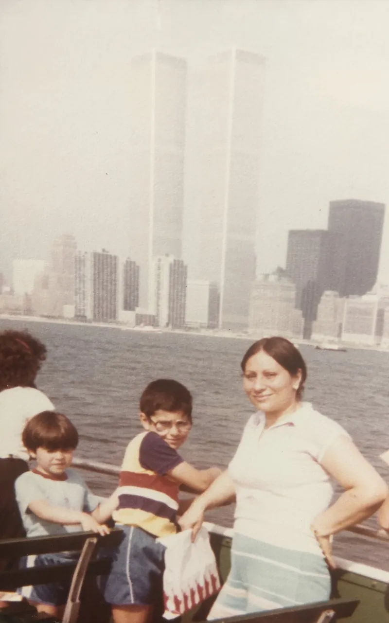 A boy with glasses, his younger brother and their mother at the rail of a boat in New York Harbor, the Twin Towers behind them